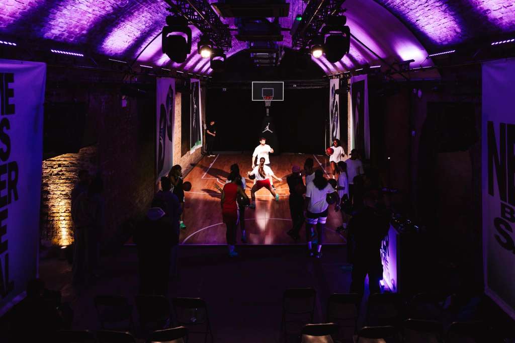 Indoor basketball court with purple ambient lighting, spectators watching players on court during active game.