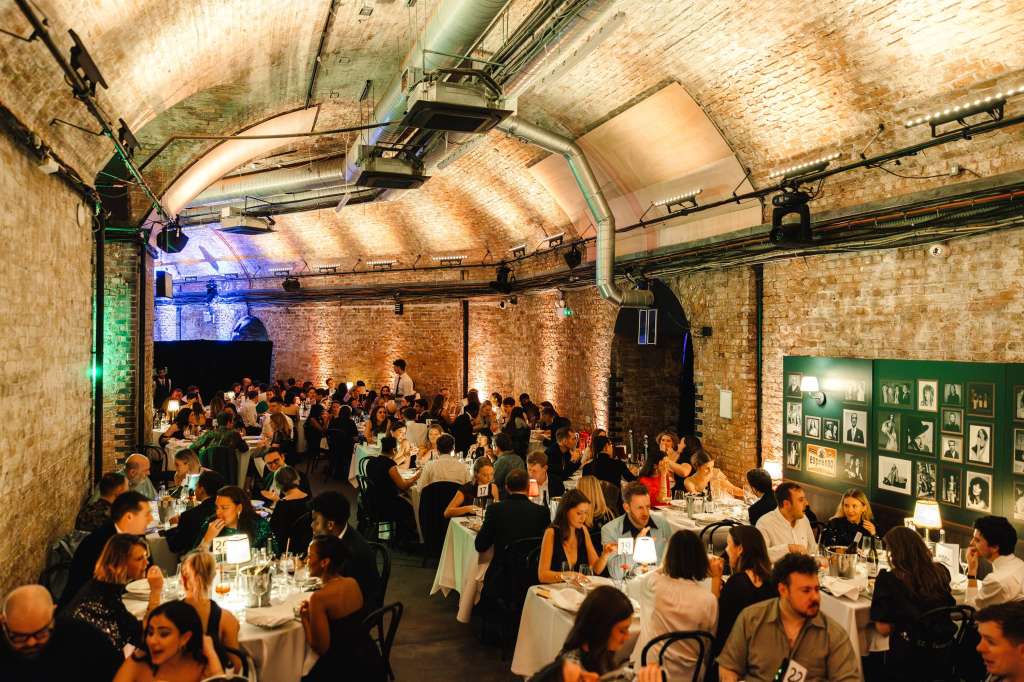 Industrial brick venue with seated guests at round tables, atmospheric lighting, and gallery wall display.