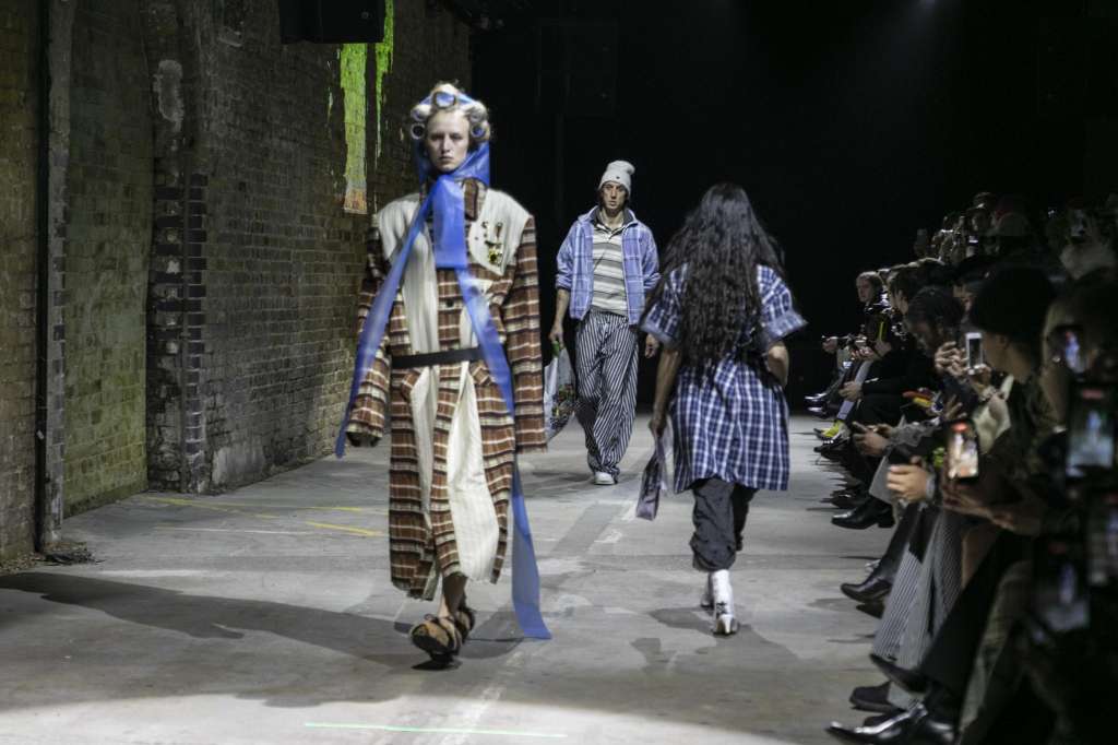 Fashion models walking down an industrial brick alley at night with seated audience members on both sides.