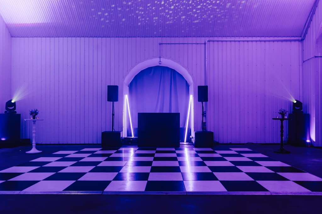 Industrial space with purple ambient lighting, checkered dance floor, neon arch, and stage setup.