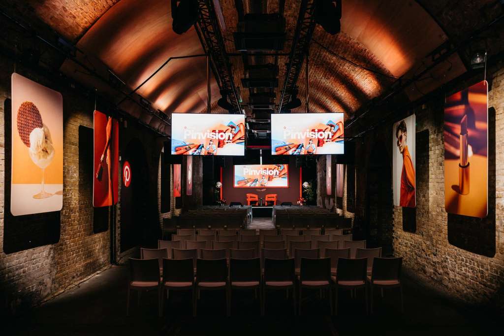 Industrial brick venue with tiered seating, stage, projection screens, and orange ambient lighting.