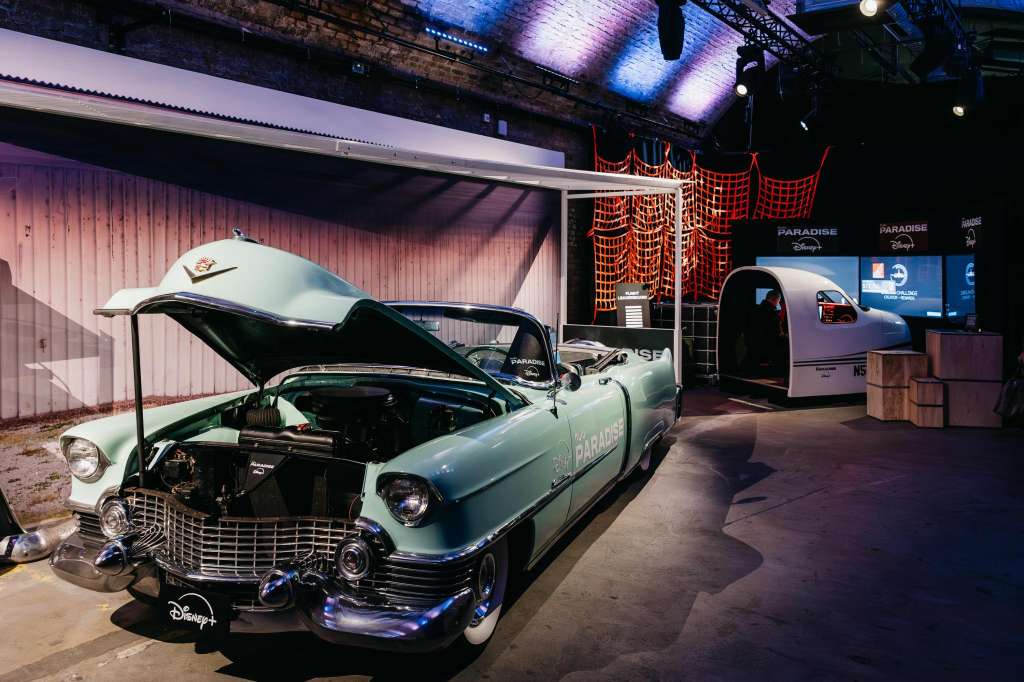 Industrial warehouse space with vintage car display, red netting, blue uplighting, and branded signage.