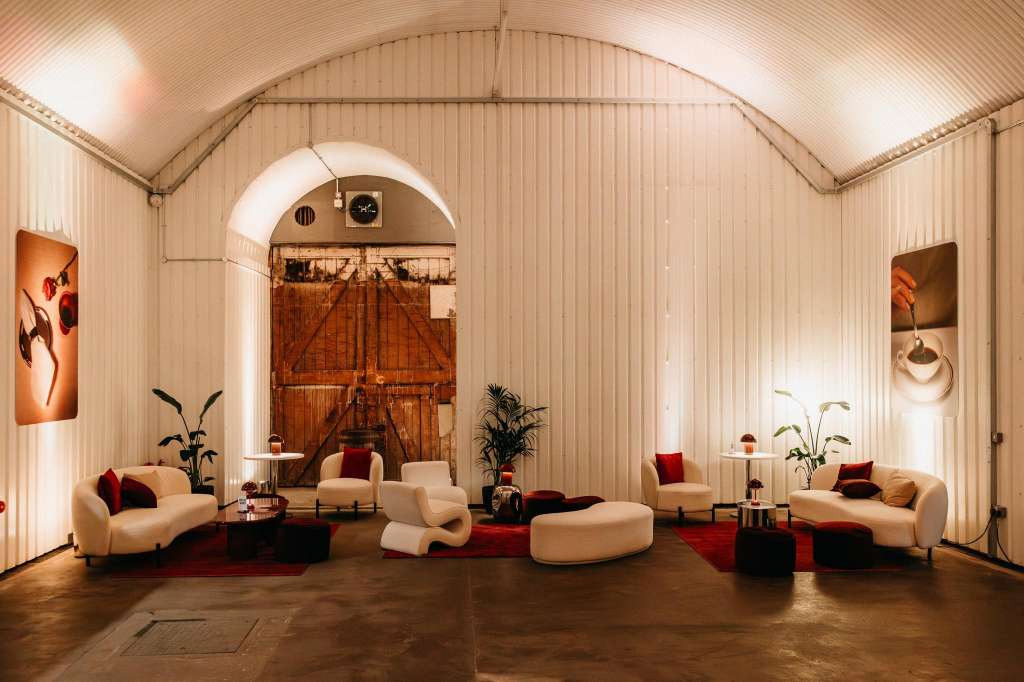 Industrial warehouse space with arched ceiling, cream curved seating, red accents, and contemporary artwork on walls.