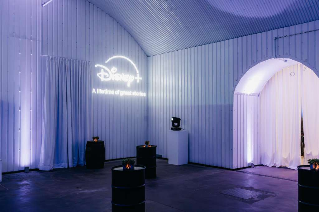 Industrial warehouse space with neon Disney signage, illuminated white fabric installations, and black sculptural elements.