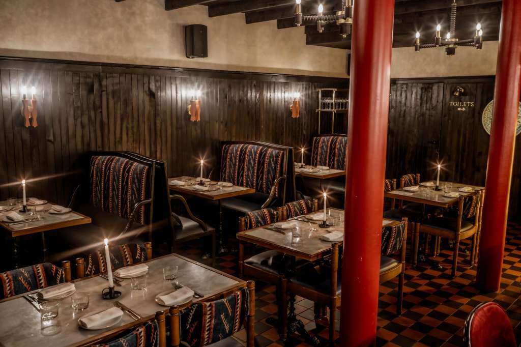 Dining room with booth seating, patterned chairs, candlelit tables, dark wood paneling, and red columns.