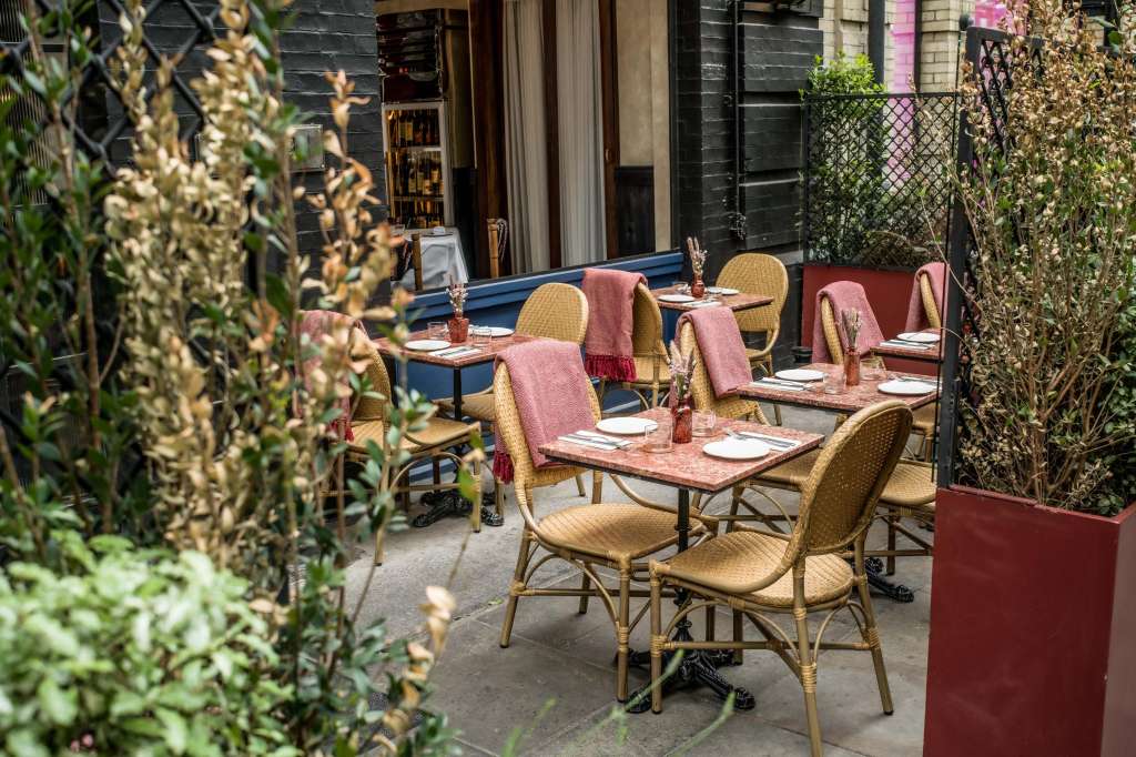 Outdoor courtyard with dining tables, woven chairs, place settings, and dried foliage in planters.
