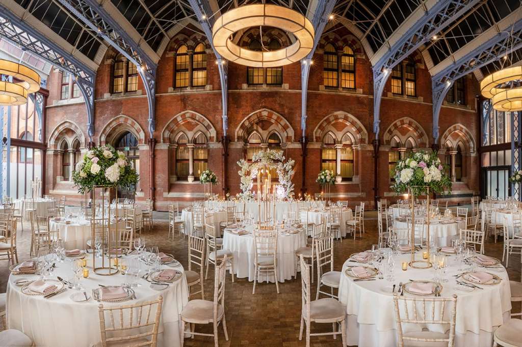 Grand hall with round tables set for dinner, white linens, floral centerpieces, and ornate brick architecture with industrial ceiling.