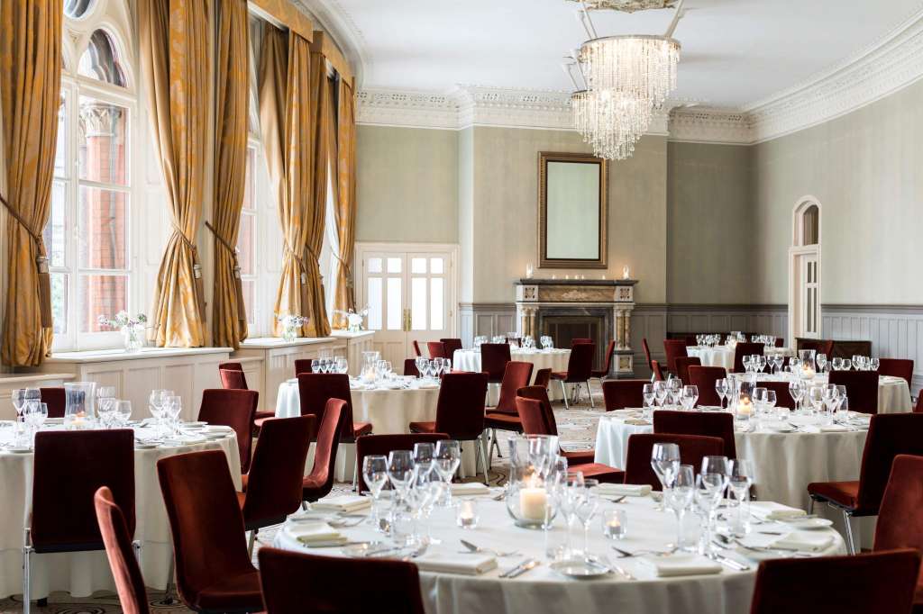 Elegant dining room with round tables, white linens, burgundy chairs, crystal chandelier, and gold curtains.