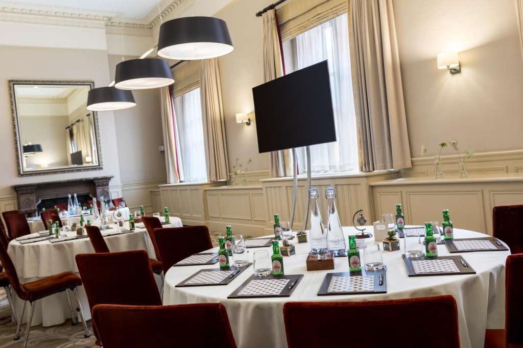 Elegant meeting room with round tables dressed in white linens, burgundy chairs, modern pendant lighting, and wall-mounted screen.