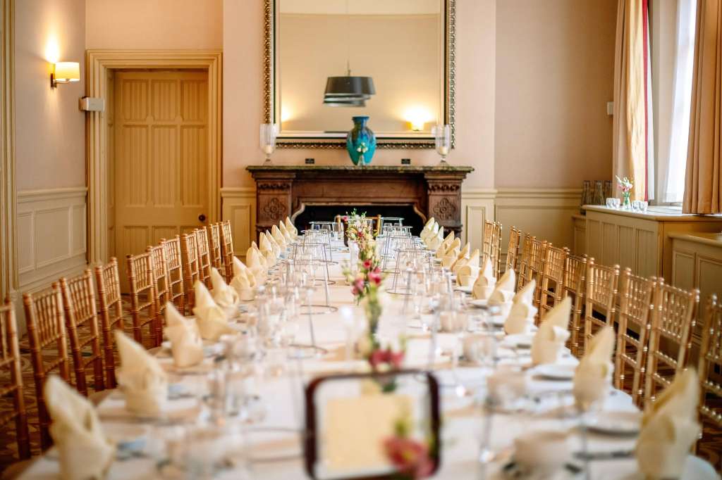 Elegant dining room with long banquet table, gold chiavari chairs, white napkins, and ornate fireplace.