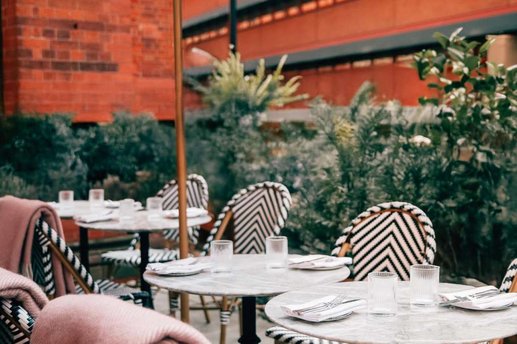 Outdoor terrace with round tables, striped chairs, umbrellas, and potted plants against red brick building.