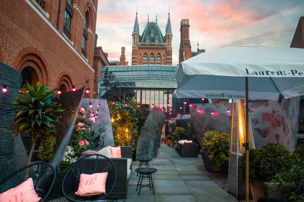 Rooftop terrace with pink string lights, potted flowers, wicker seating, and umbrellas overlooking historic architecture.