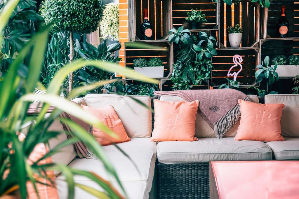 Outdoor lounge seating with coral cushions, potted plants, and neon flamingo sign against wooden slat backdrop.