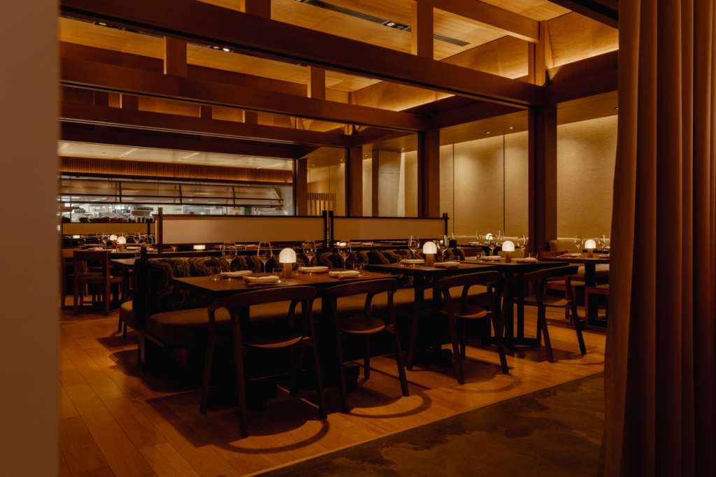 Restaurant dining room with wooden tables, modern chairs, warm ambient lighting, and open kitchen view.