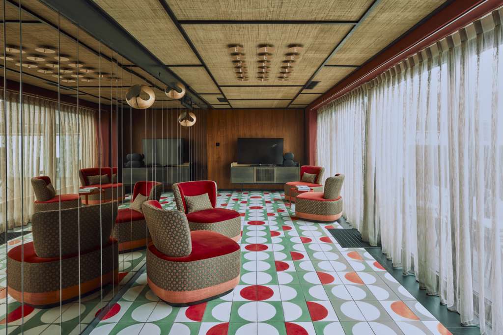 Modern lounge with red and beige armchairs, geometric floor tiles, wooden ceiling, and floor-to-ceiling windows with sheer curtains.