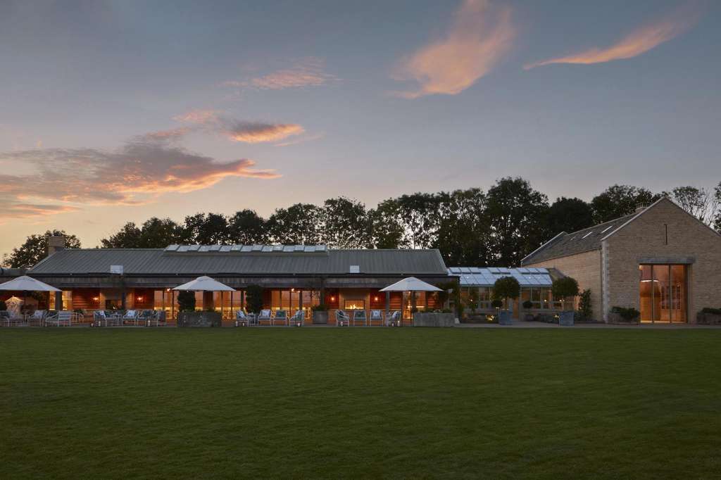 Modern stone and timber venue buildings with lit interiors at dusk, viewed across open lawn with trees beyond.