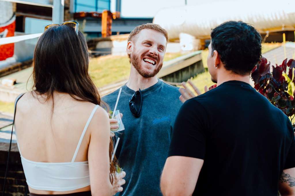 Three people socializing outdoors at an industrial rooftop venue with metal structures and green space visible.