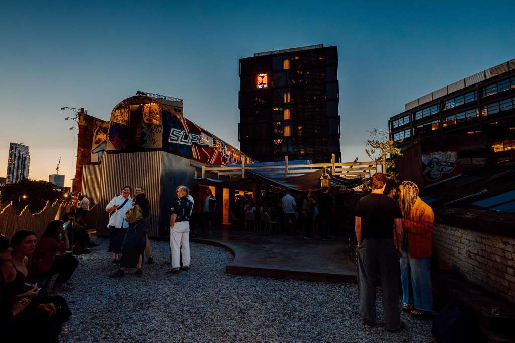 Outdoor rooftop venue at dusk with industrial shipping containers, string lighting, and guests mingling in casual groups.