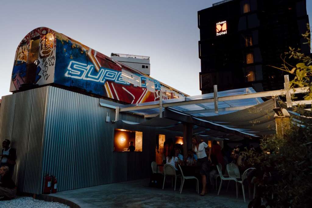 Exterior courtyard at dusk with vintage airplane fuselage, industrial structure, and guests mingling under warm lighting.