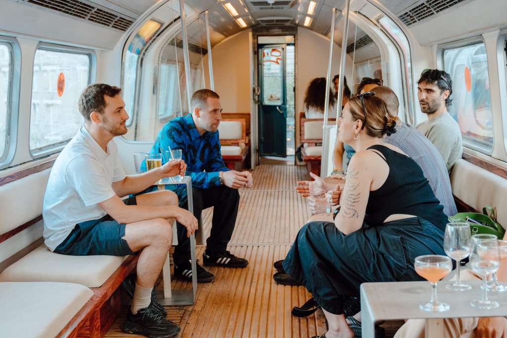 Interior of a boat or vessel with six people seated on benches, holding drinks and conversing.