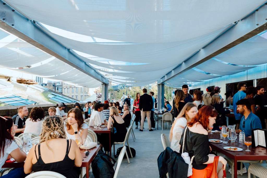 Outdoor rooftop space with white shade sails, guests seated at tables and standing, summer daylight.