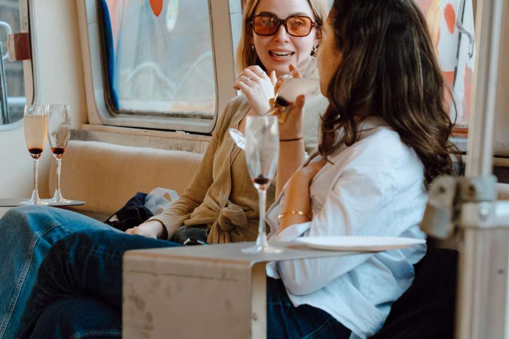 Two women seated in a boat cabin with champagne glasses and large windows overlooking water.