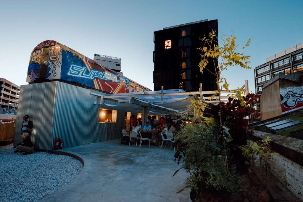 Outdoor rooftop venue with industrial corrugated structure, string lighting, and guests seated at tables under clear sky.