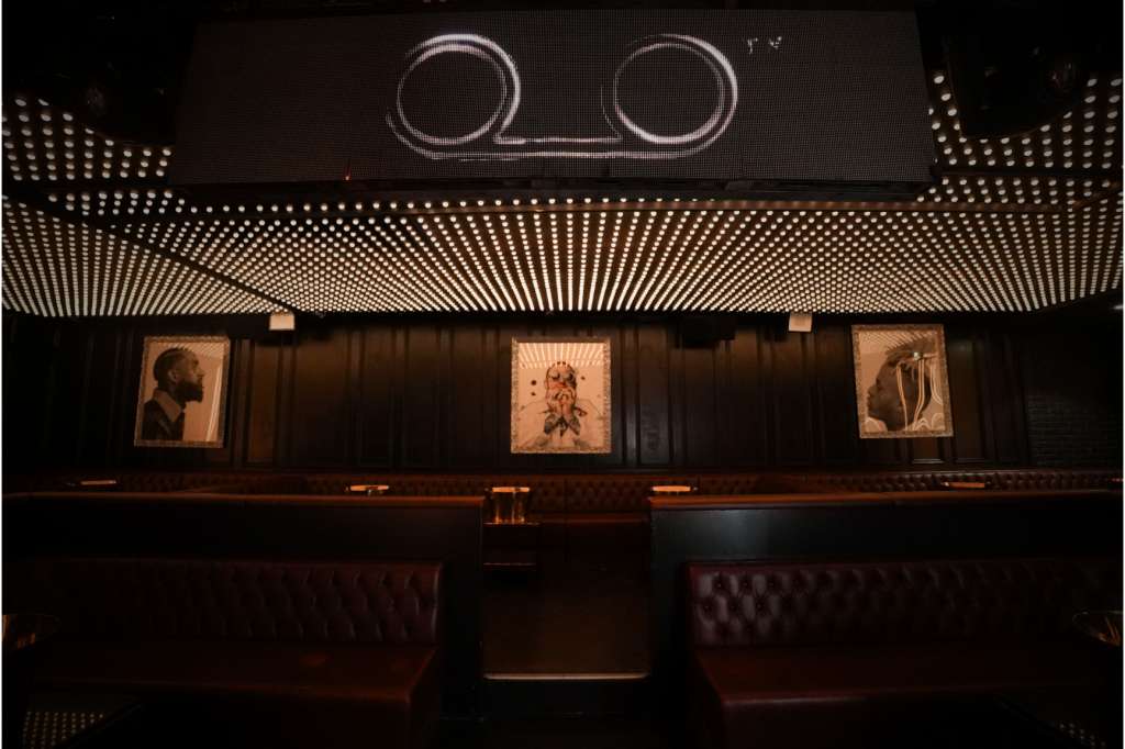 Upscale bar interior with leather seating, dark wood paneling, framed artwork, and geometric ceiling lighting.