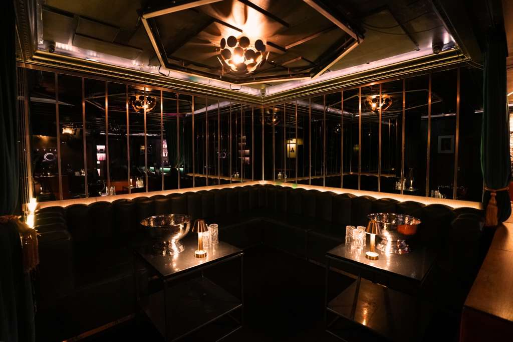 Modern lounge with black seating, low tables, candlelit ambiance, and floor-to-ceiling windows with warm lighting.