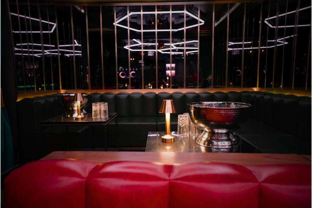 Modern lounge with red and black seating, neon geometric lighting fixtures, and floor-to-ceiling windows overlooking city lights.