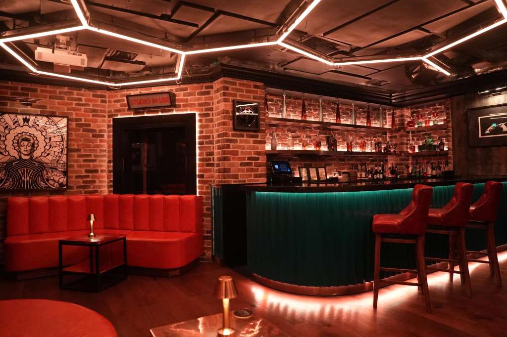 Modern bar lounge with red velvet seating, neon ceiling lights, exposed brick walls, and teal bar counter.