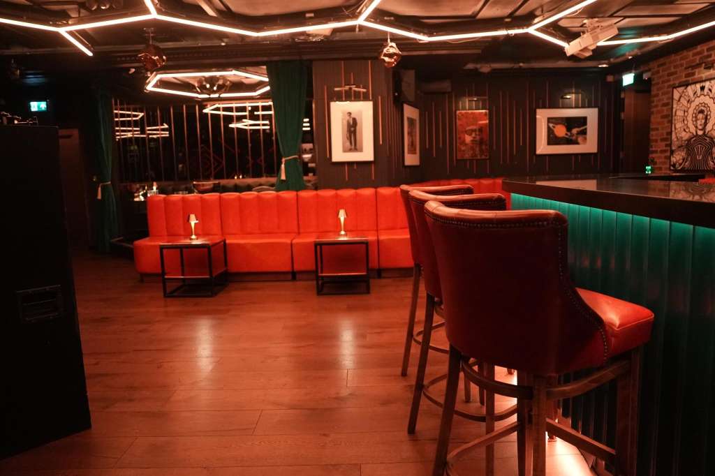 Main room with red velvet seating, neon lighting, framed artwork on dark walls, and bar counter.