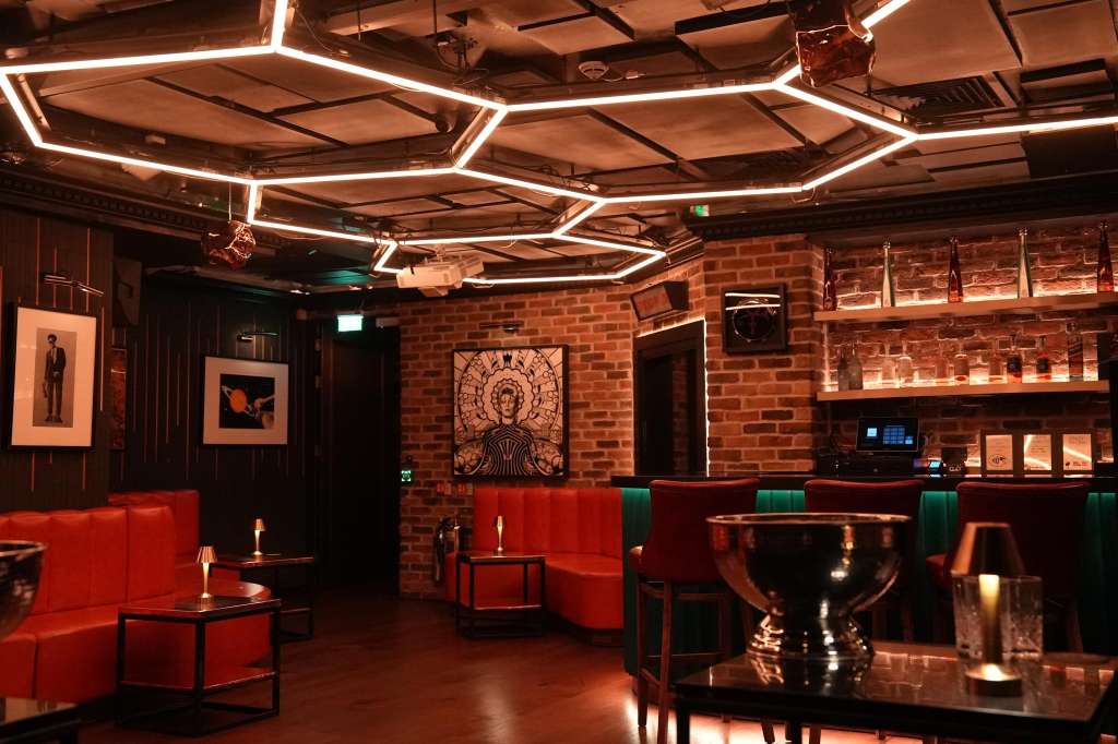 Main room with red seating, geometric neon ceiling lights, exposed brick walls, and bar shelving.