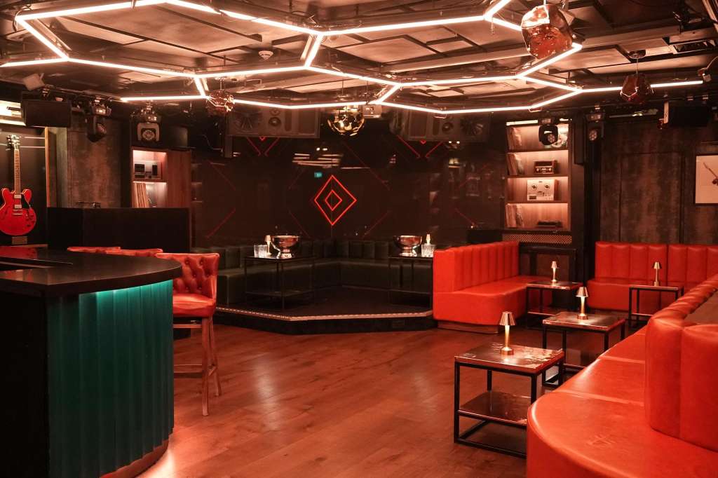 Modern event space with red velvet seating, neon ceiling lights, bar counter, and stage area with geometric backdrop.