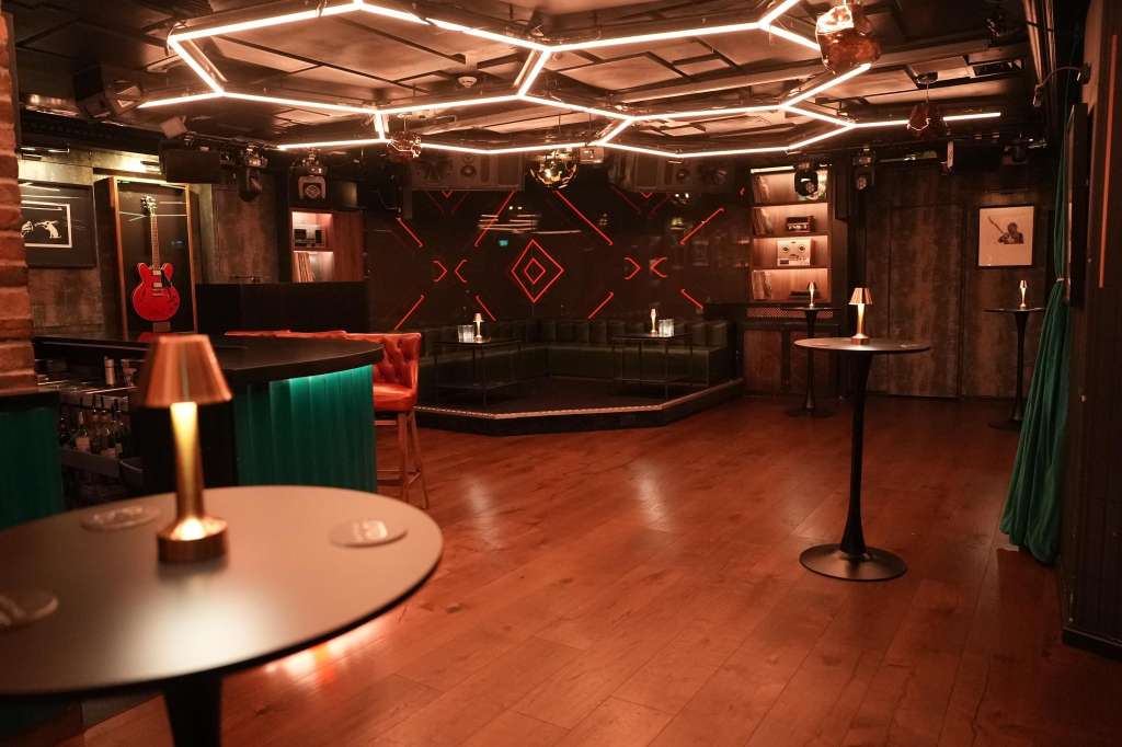 Modern event space with neon ceiling lights, bar seating, round tables, and red guitar display on dark walls.