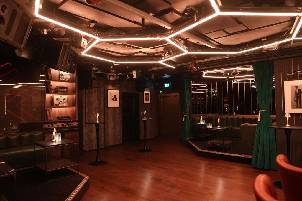 Modern event space with neon ceiling lights, dark wood paneling, bar seating, and green velvet drapes.
