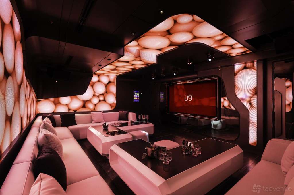 Modern karaoke lounge with pink seating, illuminated bubble wall panels, large screen displaying U9 branding, and black tables.