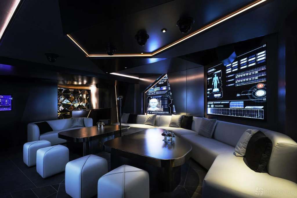 Modern lounge with curved seating, dark tables, LED screens on walls, and ambient blue lighting throughout.