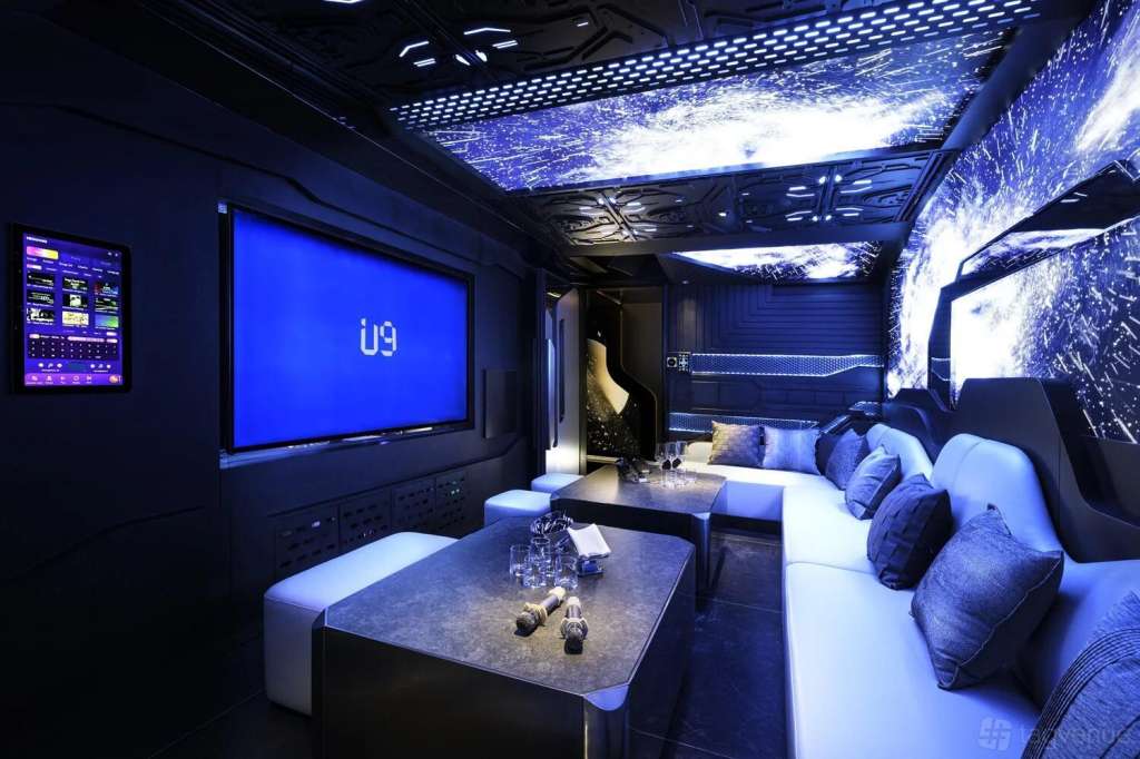 Modern karaoke lounge with blue ambient lighting, large screen displaying U9 logo, white seating, and illuminated ceiling panels.