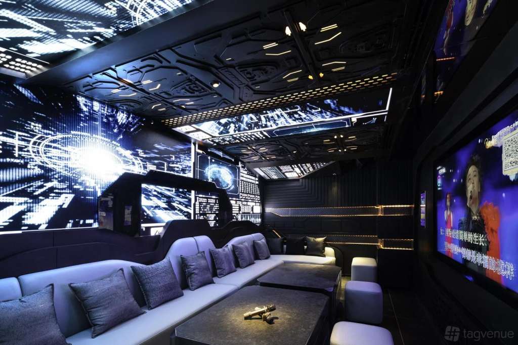 Modern karaoke lounge with LED screens, blue ambient lighting, white seating, and dark ceiling with geometric patterns.
