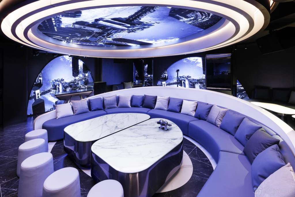 Modern lounge with curved blue seating, marble tables, neon ceiling lights, and panoramic city views on screens.