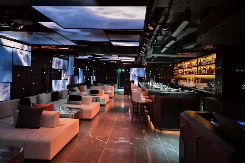 Modern cocktail lounge with modular seating, illuminated bar, video screens, and ambient lighting.