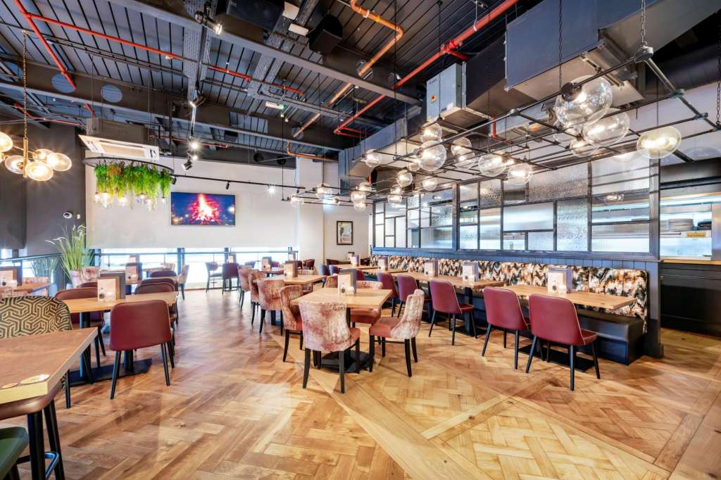 Modern dining space with wooden tables, burgundy chairs, exposed ceiling with red neon and pendant lighting, and floor-to-ceiling windows.