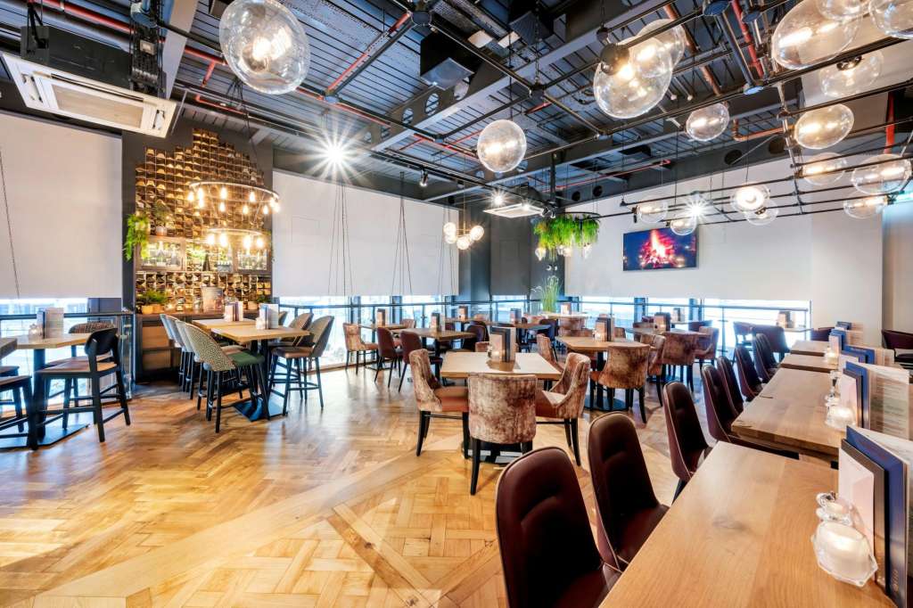 Modern dining space with mixed seating, pendant lighting, exposed ceiling, and illuminated bar wall.