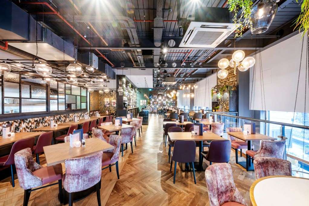Modern restaurant interior with wooden tables, upholstered chairs, pendant lighting, and floor-to-ceiling windows.