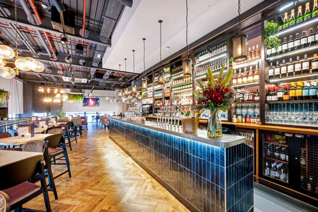 Modern bar interior with illuminated shelving, central bar counter, dining tables, and contemporary lighting fixtures.