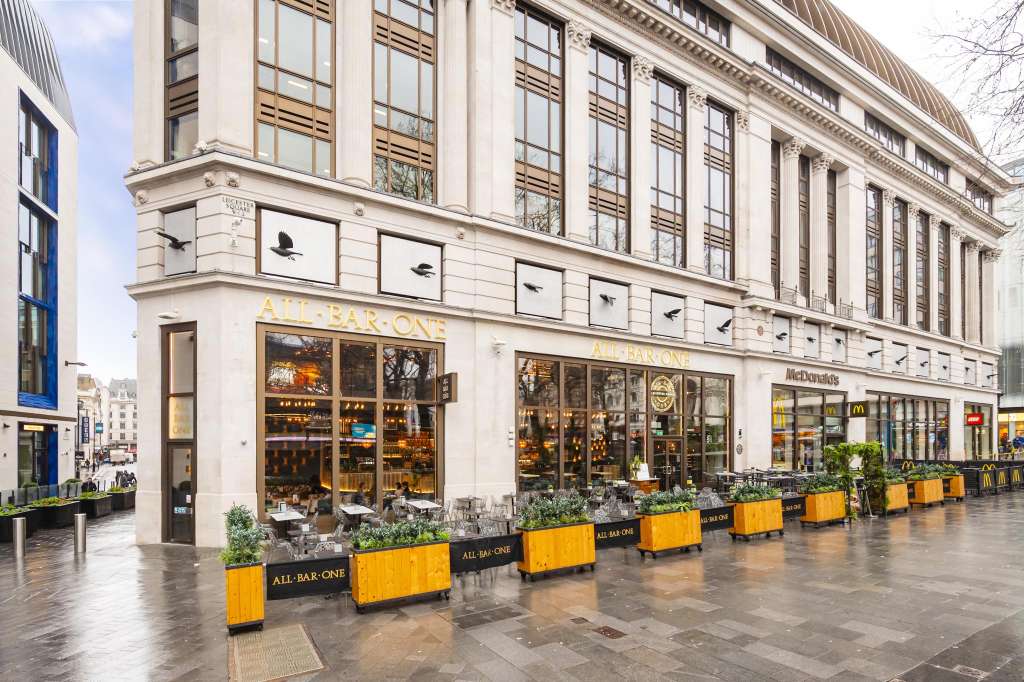 Exterior storefront of All Bar One Leicester Square with large windows, yellow planters, and classical architecture.