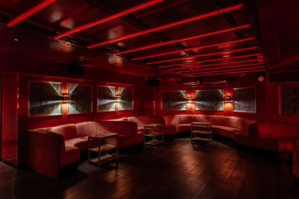 Modern lounge with red ambient lighting, linear ceiling fixtures, built-in seating, and framed artwork on walls.