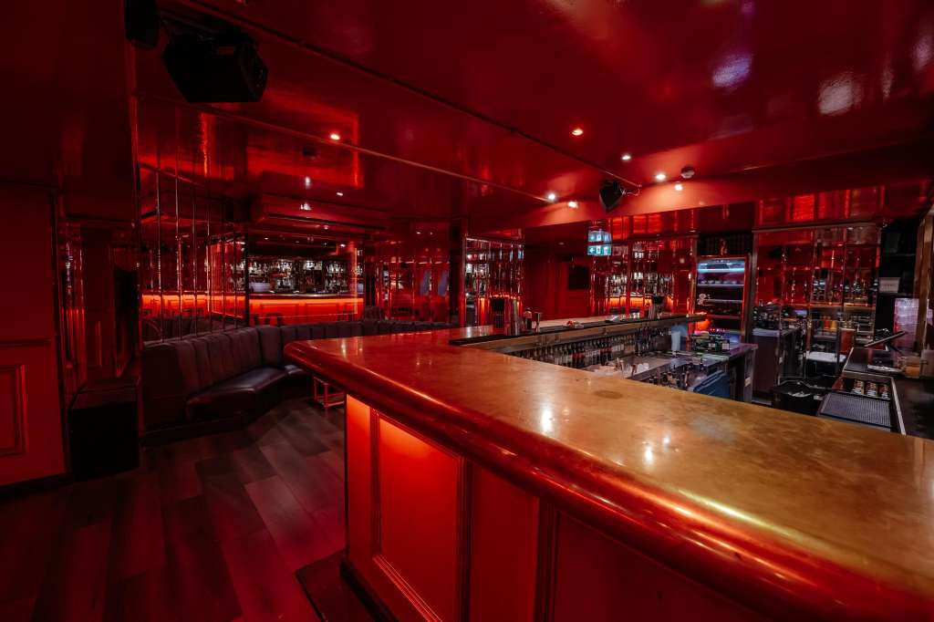 Modern bar interior with long wooden counter, red ambient lighting, shelving with bottles, and lounge seating.