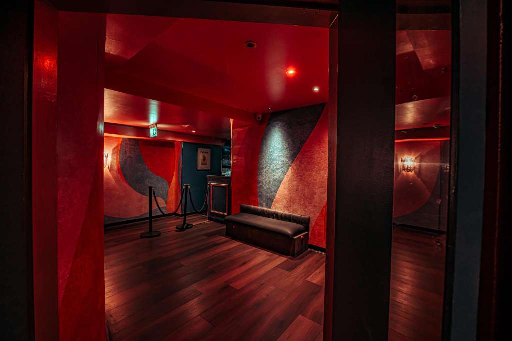 Modern interior with red-lit walls, dark wood flooring, geometric artwork, and minimal furnishings.